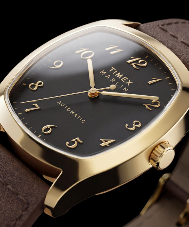 Altın tonlu Timex Marlin Chatham Automatic'in siyah kadranını, aplike altın tonlu Arap rakamlarını, altın tonlu ibrelerini ve kahverengi deri kayışını gösteren yakın çekim