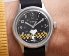 Timex MK1'in siyah kadranının yakın çekimi; sarı-siyah kareli bir arka plan üzerinde Snoopy figürü, Arap rakamlı saat işaretleri ve ışıklı ibreler görülüyor.