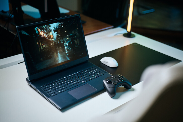 Beyaz bir stüdyo masasının üzerine yerleştirilmiş, açık renkli bir Alienware oyun dizüstü bilgisayarı, keskin yol izleme detaylarıyla karanlık, yansıtıcı bir sokak sahnesini çalıştırıyor; bilgisayarın yanında siyah bir oyun kumandası ve beyaz bir kablosuz fare bulunuyor.