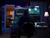 Kapüşonlu bir araştırmacı karanlık bir ortamda birden fazla ekran üzerinde çalışıyor. Jenerik hacker resimde. 