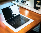 Sıcak tonlu ahşap bir masanın üzerinde, beyaz bir masa örtüsünün üzerine yerleştirilmiş, açık mat siyah renkli bir Razer Blade 16 oyun dizüstü bilgisayarı, canlı siyah-beyaz bir duvar kağıdı görüntülüyor.