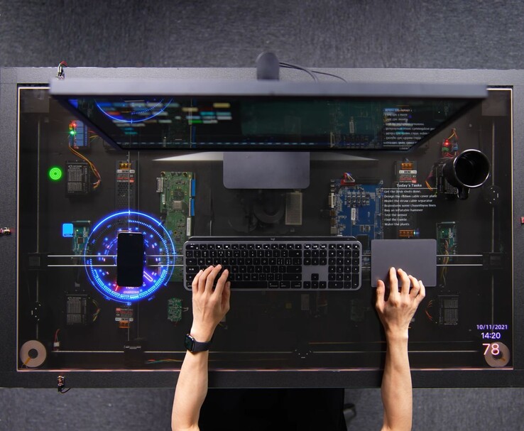 HTX Studio’nun AirPower’dan ilham alan DIY masası kullanımda; şeffaf ekranda bir telefon şarj olurken, yanında klavye, izleme paneli ve monitör bulunuyor.