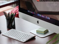 Masada duran bir iMac; ekranda macOS dock'u görünüyor ve önünde bir Apple klavyesi var