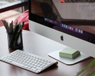 Masada duran bir iMac; ekranda macOS dock'u görünüyor ve önünde bir Apple klavyesi var