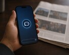 Bir el, üzerinde sohbet robotu simgesi bulunan bir akıllı telefonu tutuyor; yanında basılı bir gazete duruyor.