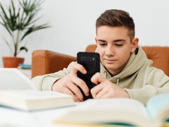 Önünde açık okul kitapları bulunan, kapüşonlu bir genç, akıllı telefonunda yazıyor.