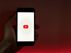 Kırmızı bir arka plan üzerinde, ekranında YouTube logosu yanan bir akıllı telefonu tutan bir el