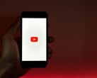 Kırmızı bir arka plan üzerinde, ekranında YouTube logosu yanan bir akıllı telefonu tutan bir el