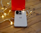 Sıcak renkli ışık zincirleriyle çevrili ahşap bir masanın üzerinde bir OnePlus 15 duruyor; arka planda ise parlak kırmızı bir OnePlus satış kutusu yer alıyor.