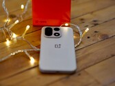 Sıcak renkli ışık zincirleriyle çevrili ahşap bir masanın üzerinde bir OnePlus 15 duruyor; arka planda ise parlak kırmızı bir OnePlus satış kutusu yer alıyor.