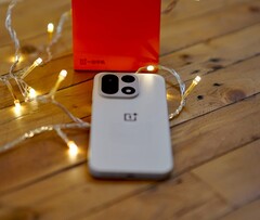 Sıcak renkli ışık zincirleriyle çevrili ahşap bir masanın üzerinde bir OnePlus 15 duruyor; arka planda ise parlak kırmızı bir OnePlus satış kutusu yer alıyor.