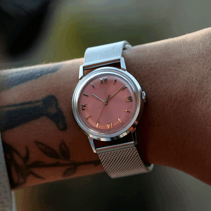 Somon rengi güneş ışını desenli kadranı ve file bileziği olan Timex x Todd Snyder Marlin Mesh saatini takan kişi.