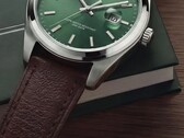 Timex Legacy Day and Date saatin koyu yeşil bir kutu ve ahşap yüzey önünde yeşil güneş ışını desenli kadranını, gümüş tonlu bezeli ve fırçalanmış kordon kulaklarını, tırtıklı kurma kolunu ve dokulu kahverengi deri kayışını gösteren yakın çekimi.