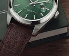 Timex Legacy Day and Date saatin koyu yeşil bir kutu ve ahşap yüzey önünde yeşil güneş ışını desenli kadranını, gümüş tonlu bezeli ve fırçalanmış kordon kulaklarını, tırtıklı kurma kolunu ve dokulu kahverengi deri kayışını gösteren yakın çekimi.