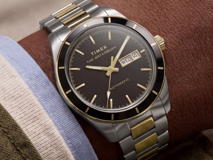 Siyah kadranlı, iki tonlu Timex Waterbury Heritage Automatic saat, bilekte takılı halde, gün-tarih göstergesini ve altın rengi detaylarını sergiliyor.
