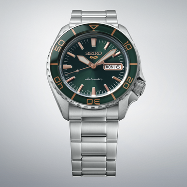 Açık gri bir arka plan önünde gösterilen, koyu yeşil kadranlı, uyumlu yeşil bezelli, pembe altın renkli ibre ve indekslere ve paslanmaz çelik bileziğe sahip Seiko 5 Sports SKX HDB005.