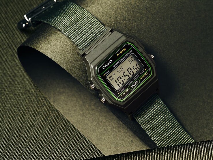 Yeşil detaylı siyah kasalı ve dokuma yeşil kumaş kayışlı Casio F-91WF-3A dijital saat, koyu yeşil dokulu bir arka plan önünde fotoğraflanmıştır.