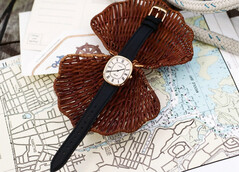 Deniz haritaları ve hasır örgü üzerinde fotoğraflanmış, beyaz Romen rakamlı kadranı ve siyah deri kayışı olan altın tonlu Noah × Timex Boatswain saat.