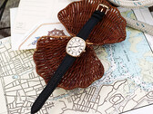 Deniz haritaları ve hasır örgü üzerinde fotoğraflanmış, beyaz Romen rakamlı kadranı ve siyah deri kayışı olan altın tonlu Noah × Timex Boatswain saat.