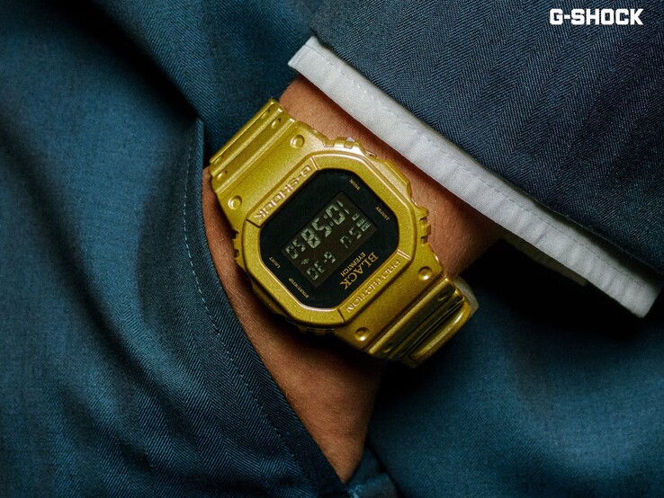 Koyu mavi kumaşın önünde, bileğe takılı altın renkli Casio G-Shock DW-5600BEP-9 saatinin yakın çekimi. Saat, siyah dijital ekrana, altın renkli kasaya ve kayışa sahiptir; çerçeve ve kadranında BlackEyePatch markası bulunmaktadır.